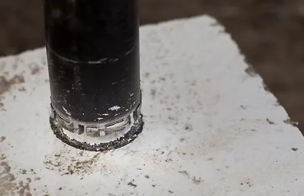 Close-up of a hole saw drill bit cutting through a white material, with dust and debris around the partially cut hole—showcasing the precision of a concrete contractor at work.