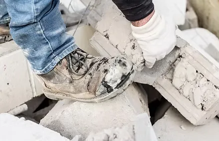 A concrete contractor wearing gloves and work boots moves a concrete block among a pile of construction rubble.