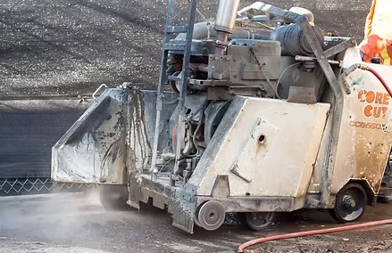 A concrete contractor operates a cutting machine, releasing dust as it slices through pavement at a bustling construction site.