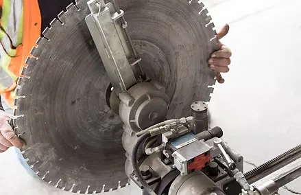 A concrete contractor holds a large industrial circular saw blade attached to a machine, wearing a safety vest.